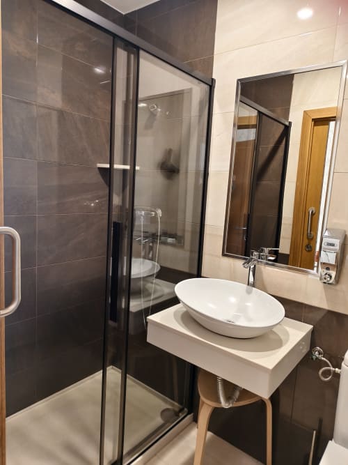 Baño moderno del albergue Ultreia en Arzúa con lavabo blanco de cerámica, ducha con azulejos oscuros y espejo iluminado