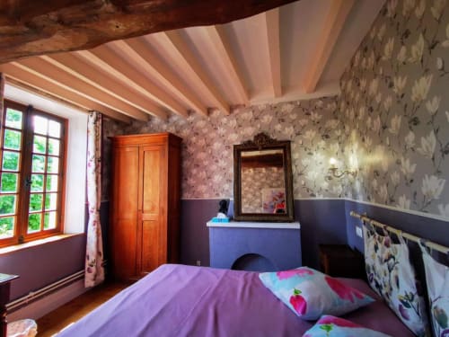 Chambre douillette au Coin des Artistes avec linge de lit violet, poutres apparentes, papier peint fleuri et vue sur le jardin