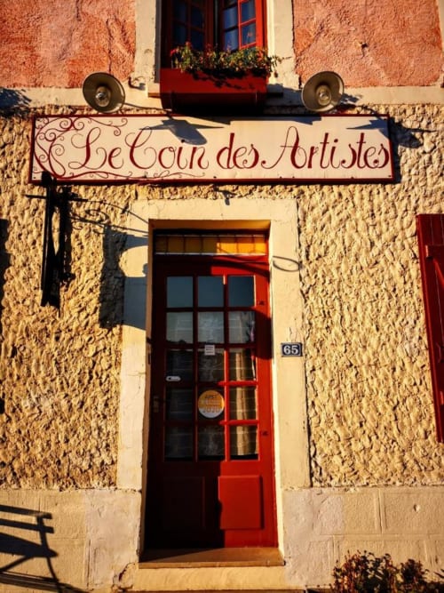 Entrée du Gîte Le Coin des Artistes avec porte rouge, enseigne ornée et façade en pierre rustique