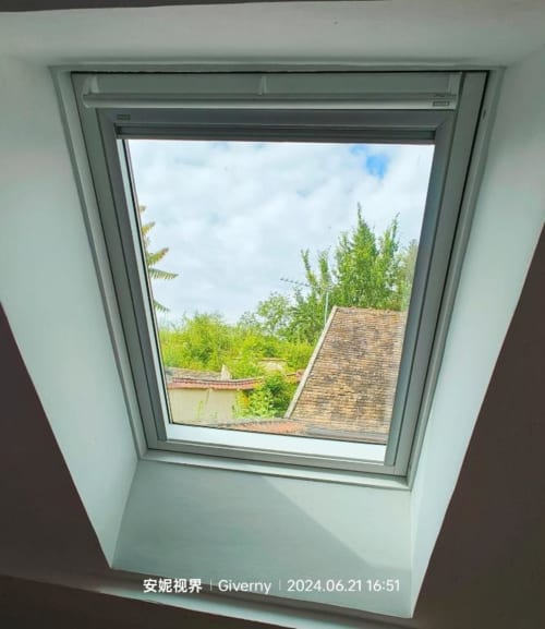 Fenêtre de toit dans la chambre mansardée du Coin des Artistes avec vue sur le jardin, les arbres et la toiture en terre cuite