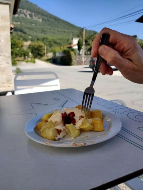 Hand holding fork over plate of potatoes with sauce at Posada de Ollo, mountain village backdrop