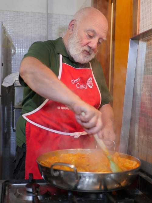 Homme chauve barbu cuisinant en tablier rouge à la cuisine du Bresbot B&B, remuant une poêle avec une sauce dorée