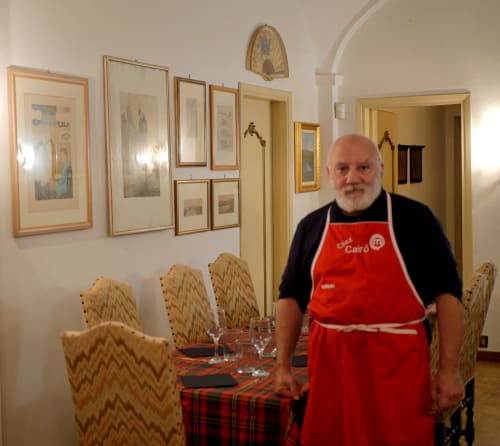 Propriétaire en tablier rouge dans la salle à manger du Bresbot B&B à Bressana, avec œuvres encadrées et chaises rembourrées