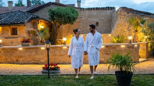 Couple in robes walking at dusk at Le Clos Des Anges B&B in Bagnols, France, with stone architecture and garden lighting.