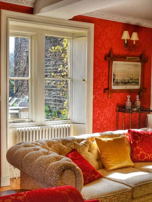 Luxuszimmer mit roten Damastmauern, cremefarbenem Chesterfield-Sofa und Gartenblick.
