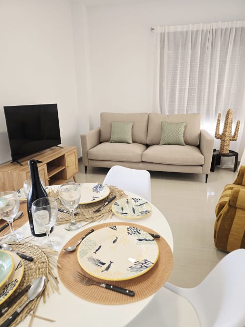 Amplia sala de estar y comedor integrados en apartamento Ona Vacaciones Gandía con sofá beige, mesa blanca y jardín