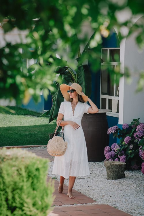 Mulher em vestido branco e chapéu de sol na boutique hotel Herdade da Cova da Onça em Canha, Portugal, rodeada por flores roxas