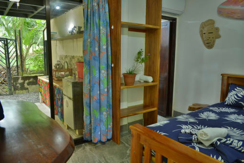 Habitación del Hotel Swell Pavones con cama de madera, divisor de cortina azul estampada, estantería, planta en maceta y vista al jardín