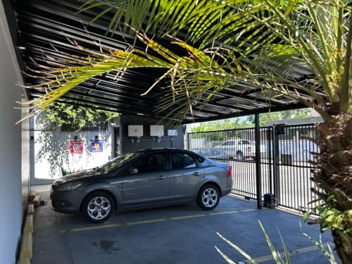 Sedã azul estacionado sob carporte coberto com portão de metal preto e plantas tropicais na hospedagem BORA BORA HOSPEDAGENS