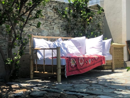 Lit rustique aux oreillers blancs et couverture à motifs rouges dans la cour ensoleillée de la villa La Conciergerie du Cap Corse, Centuri