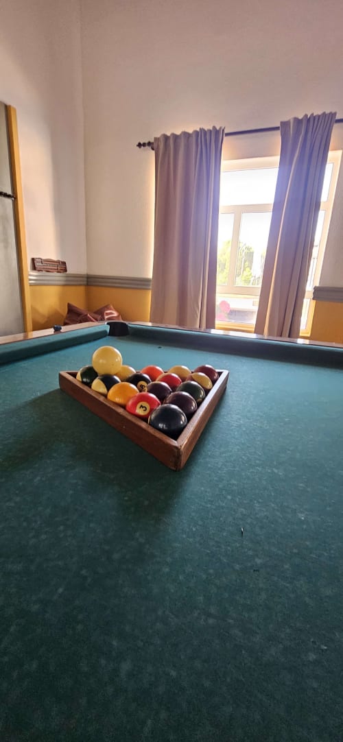 Mesa de bilhar com bolas de sinuca em suporte de madeira na casa rural Herdade da Cova da Onça, cortinas roxas e vista para o jardim