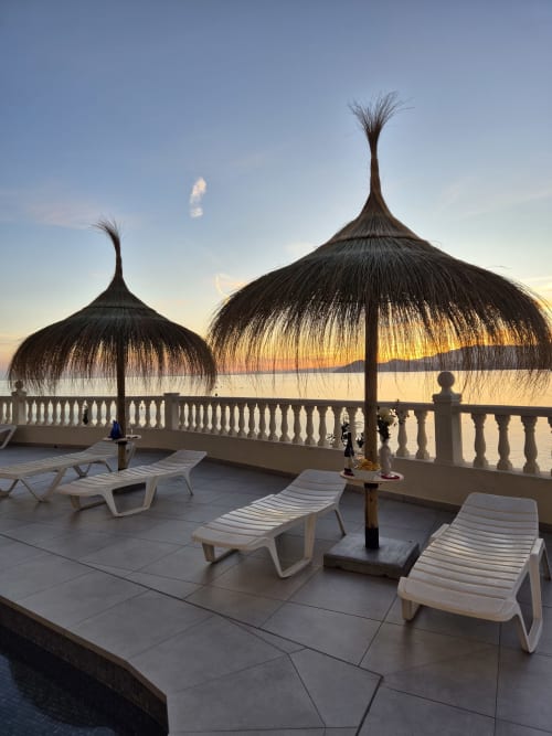 Terraza frente al mar en Portofino Playa con sombrillas de paja, tumbonas y vistas al atardecer