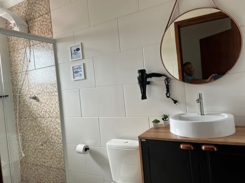 Banheiro moderno do apartamento BORA BORA com espelho redondo, pia branca, vaso sanitário e chuveiro revestido em azulejos na Costa e Silva, Joinville