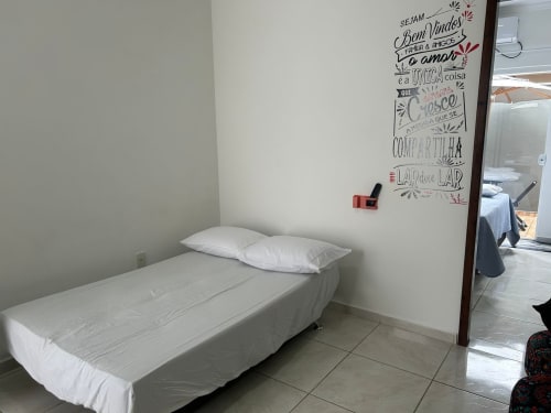 Quarto simples em apartamento de Joinville com cama branca, piso de cerâmica e arte de parede motivacional portuguesa no BORA BORA