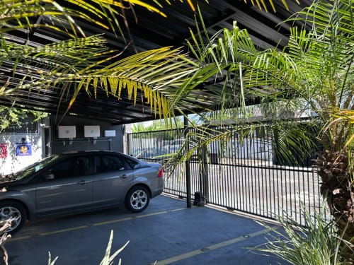 Garagem coberta com sedã azul no apartamento BORA BORA HOSPEDAGENS em Joinville, Brasil, com plantas de palmeira e segurança em preto