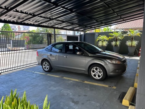 Sedan cinza estacionado na garagem coberta do apartamento BORA BORA, Joinville, com portão preto e plantas em vasos à vista.
