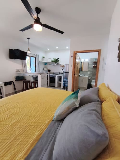 Ropa de cama amarilla con almohadas en el estudio Diosa del Mar, cocina abierta y baño visible, ventilador de techo