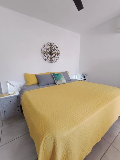 Dormitorio moderno con edredón acolchado amarillo, cojines decorativos gris y azul verdoso, ornamentación elegante en pared, mesita de noche y techo blanco