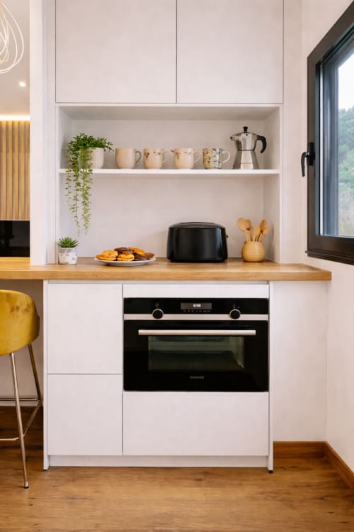 Cocina moderna blanca con horno integrado, encimera de madera, estantería abierta con tazas y plantas en apartamento de Baiona