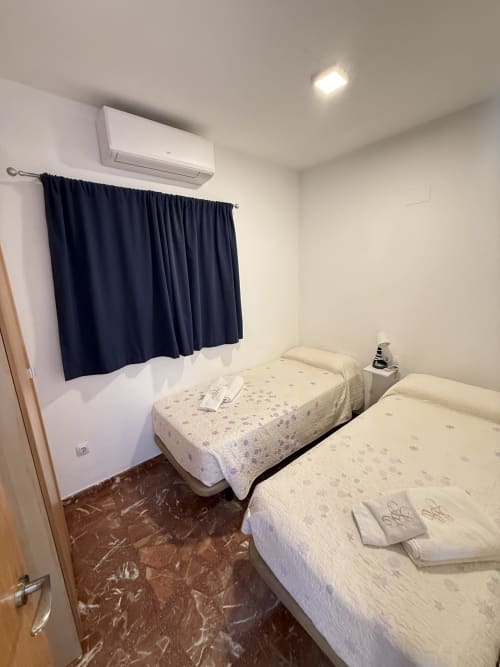Dormitorio luminoso con dos camas individuales, cortinas azul oscuro, aire acondicionado y piso de mármol en apartamento de Matalascañas
