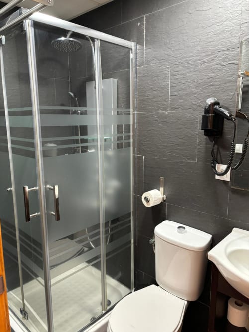 Baño moderno con paredes de azulejos oscuros, ducha de cristal, inodoro blanco y secador de pelo en apartamento de Matalascañas