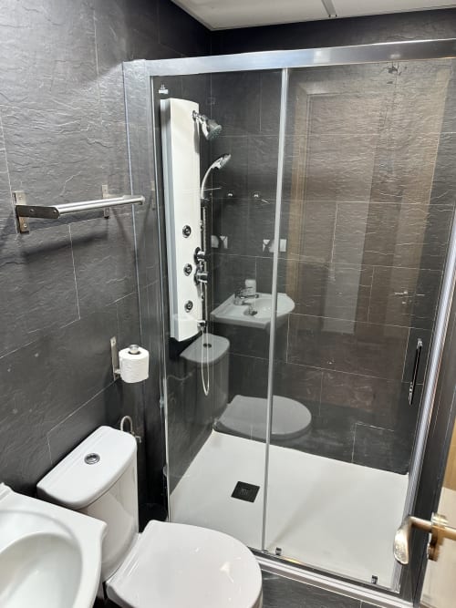 Baño moderno con paredes de azulejos negros, cabina de ducha de vidrio, inodoro blanco y lavabo en apartamento de Apartamentos A la