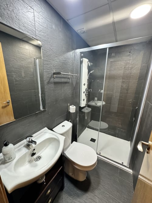 Baño moderno en apartamento frente al mar con paredes de azulejos oscuros, lavamanos blanco, inodoro y ducha a ras de piso con cabina de cristal