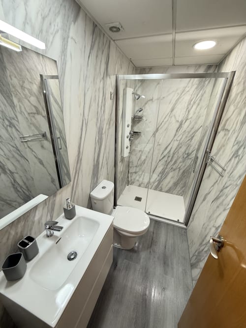 Baño moderno con azulejos efecto mármol, lavabo blanco, inodoro y cabina de ducha de cristal en Apartamentos A la Orilla del