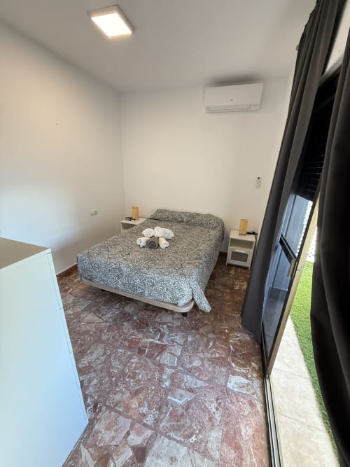 Dormitorio con cama de patrón gris, mesillas blancas, piso de baldosas, aire acondicionado y puerta de cristal hacia terraza con vistas al mar