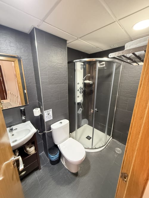 Baño moderno en apartamento de Matalascañas con paredes de azulejos oscuros, inodoro blanco, ducha de esquina, lavabo y muebles de madera