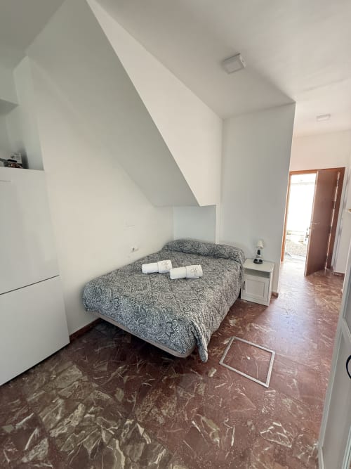 Dormitorio con ropa de cama gris estampada, paredes blancas, techo inclinado y piso de terracota en apartamento de Matalascañas.