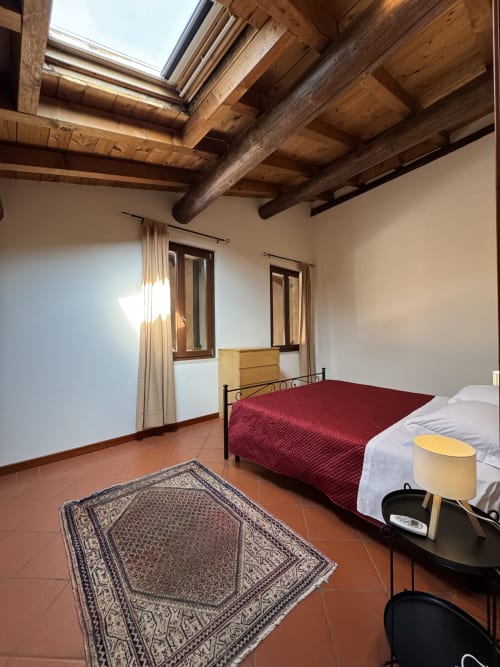 Bilocale Dalì apartment bedroom at Corte Roveggia, Verona, with exposed wooden beams, red-covered bed, and terracotta floor