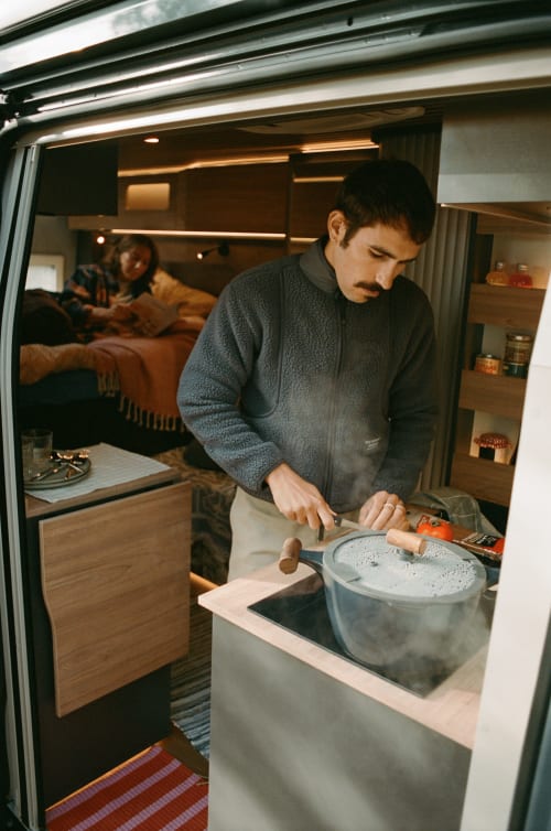 Hombre cocinando en la cocina de autocaravana La Nómada mientras pareja descansa de fondo, interior acogedor con iluminación cálida