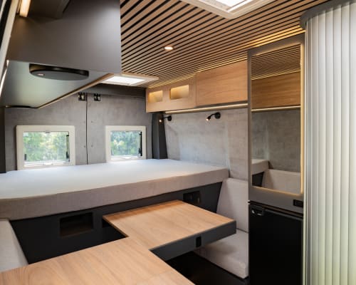 Interior moderno de autocaravana en La Nómada con cama, área de comedor, cocina y ventanales con vistas a la naturaleza.