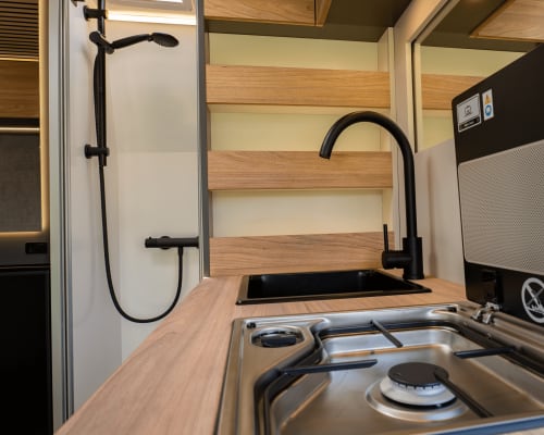 Cocina y baño modernos de autocaravana con estanterías de madera, accesorios negros, estufa de gas y ducha en La Nómada por Cien Vinos