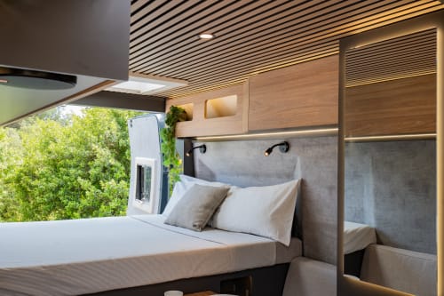 Dormitorio moderno de autocaravana con cama queen, almacenamiento de madera, cabecero gris y vista a árboles verdes en La Nómada