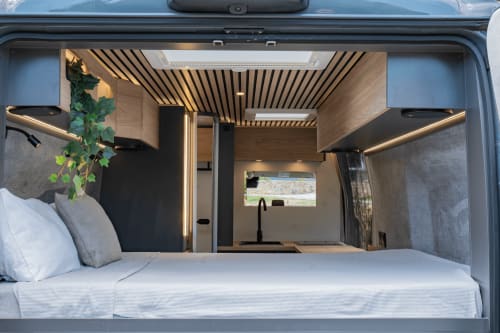 Interior de la autocaravana La Nómada Por Cien Vinos con cama matrimonial, cocina integrada, iluminación cálida y diseño moderno en madera