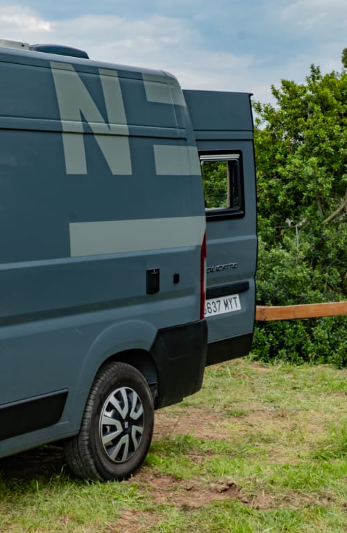 Autocaravana Ducato azul estacionada en el camping La Nómada, rodeada de vegetación verde y cerca de madera