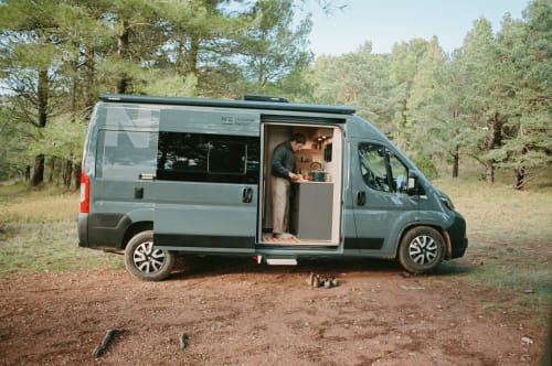 Campervan gris NEO estacionada en claro del bosque con persona adentro, alojamiento La Nómada en España