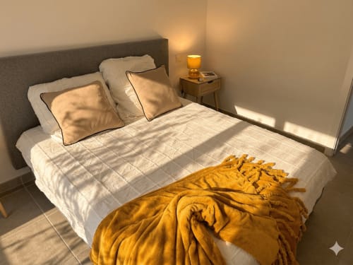 Chambre ensoleillée de La villa Métisse avec literie blanche, oreillers dorés, jeté jaune et table de nuit en bois