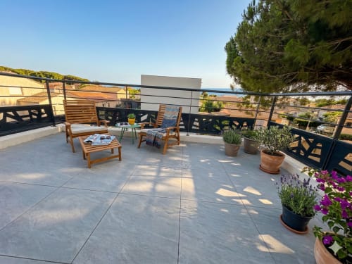 Terrasse ensoleillée de la villa Conciergerie IB avec transats en bois, fleurs en pots, vue mer et paysage méditerranéen