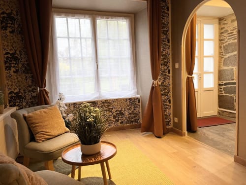 Chambre lumineuse du Manoir Des Haies avec fauteuil, table ronde, grandes fenêtres sur jardin et arcades menant à l'entrée