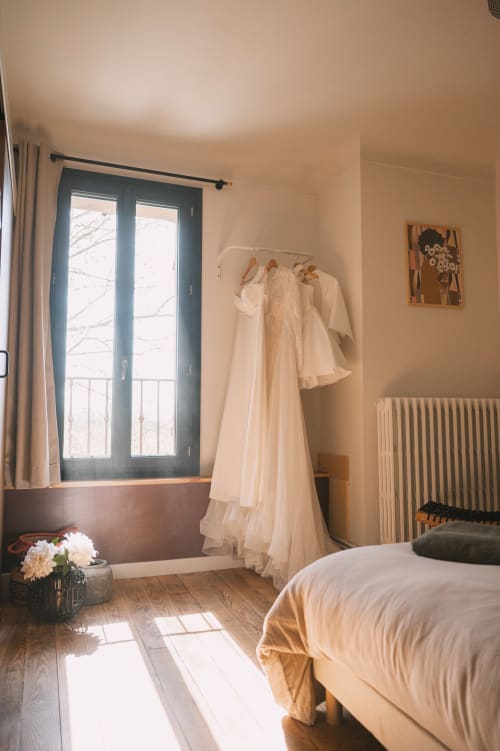 Chambre lumineuse au Mas de la Vernède à Aramon avec literie crème, portes-fenêtres et robe de mariée sur cintre.
