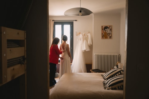 Femme en rouge aidant la mariée avec sa robe blanche dans la chambre de l'hôtel Le Mas de la Vernède à Aramon