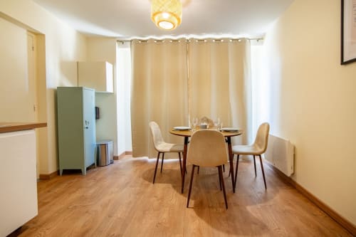 Salle à manger de studio moderne avec table ronde, chaises beige, kitchenette et rideaux avec vue sur la ville à Saint Laurent sur