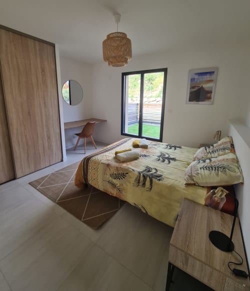 Modern bedroom in a Corsican villa with a pool view, botanical-patterned bedding, a rattan pendant light and a small workspace, managed by the Corsu di Core concierge service.