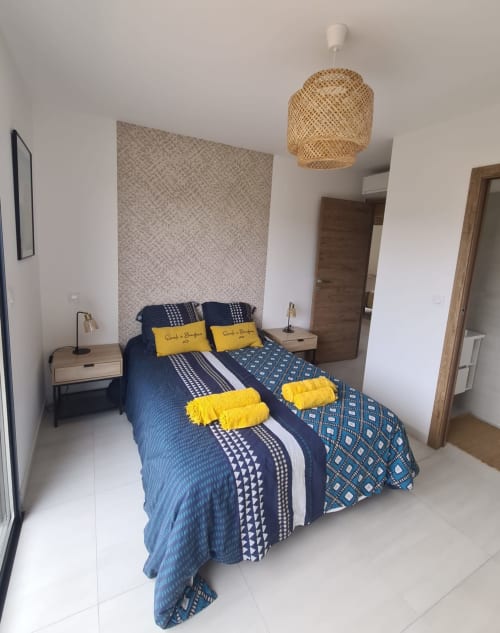 Modern bedroom in a Corsican villa featuring a patterned blue bedding set, yellow accents, a woven pendant light and a textured wall, managed by the Corsu di Core concierge service.