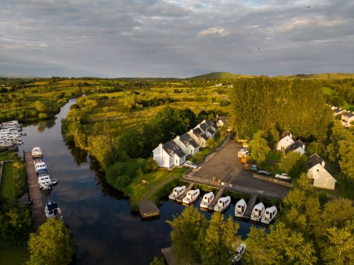Aerial view of Leitrim Quay cottages on riverbank with moored boats, garden, and countryside landscape