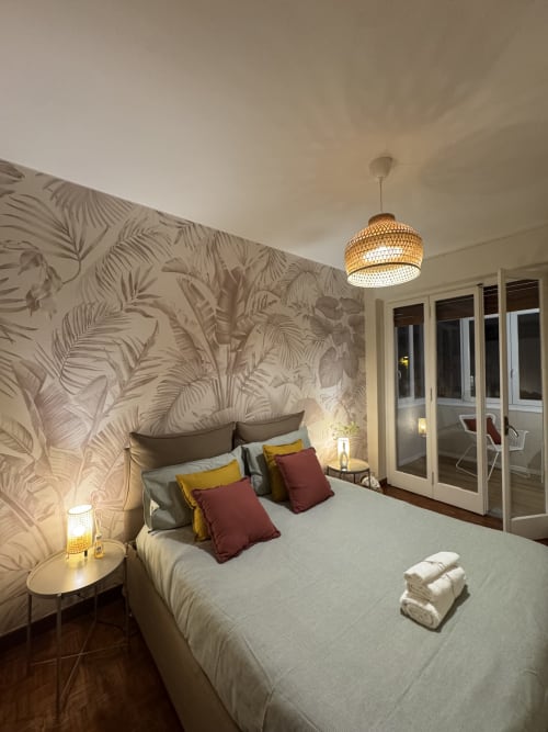 Camera moderna a Sanremo con carta da parati botanica, letto con cuscini colorati, lampada sospesa intrecciata e balcone con vista città