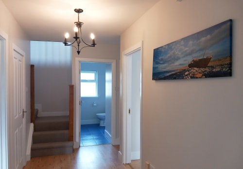 Modern hallway at Leitrim Quay cottage in Ireland with blue bathroom visible, pendant chandelier, and seascape artwork on